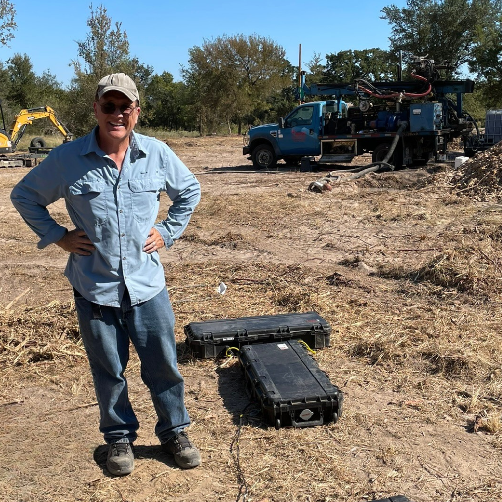 Andrew from Well Water Finders standing on a Texas property with patented groundwater detection equipment in the foreground and a blue drilling rig truck in the background — the on-site moment where survey data meets the drillers who use it.