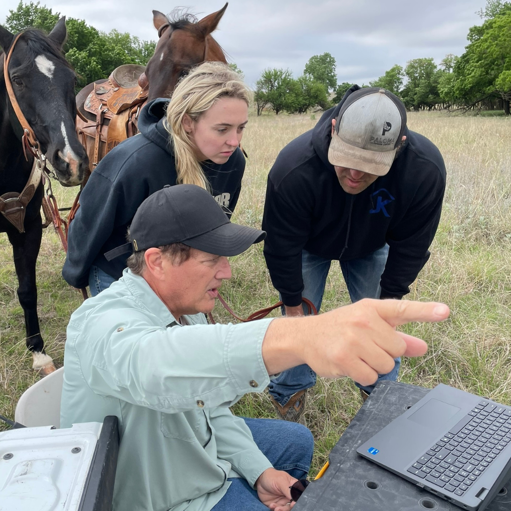 Andrew from Well Water Finders pointing to a laptop screen during an on-site groundwater survey, walking two Texas ranchers through the data while their horses graze nearby — the consultation moment after the equipment captures readings at their property.