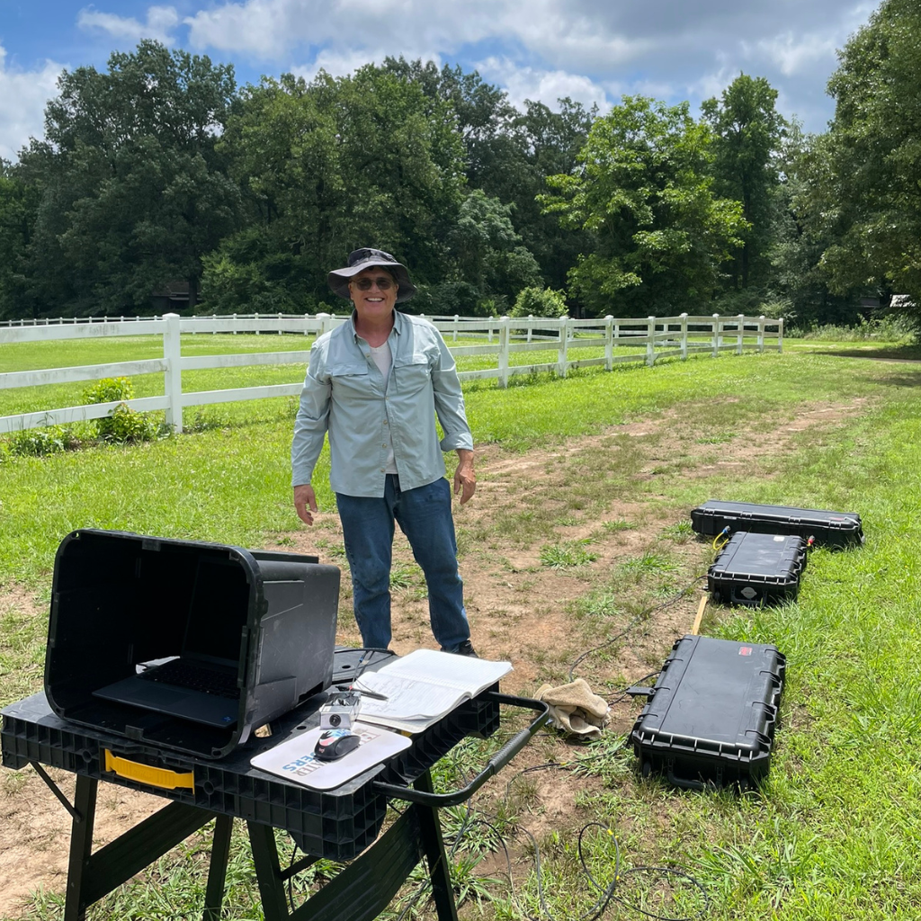 Andrew from Well Water Finders standing at the survey setup on a Texas landowner's property, with patented groundwater detection equipment cases positioned across the field and survey notes on the work table — the on-site visit that delivers point-specific groundwater detection at coordinates a driller can use.